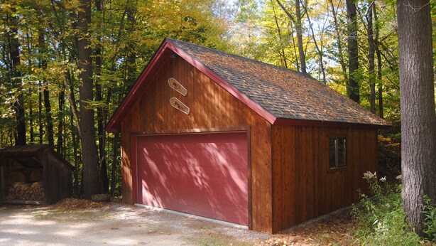 Un abri en bois avec une porte de garage rouge, entouré d’arbres.