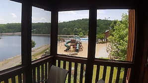 Vista desde una habitación con barandilla de madera, sobre un río, un muelle con equipos y una colina boscosa.