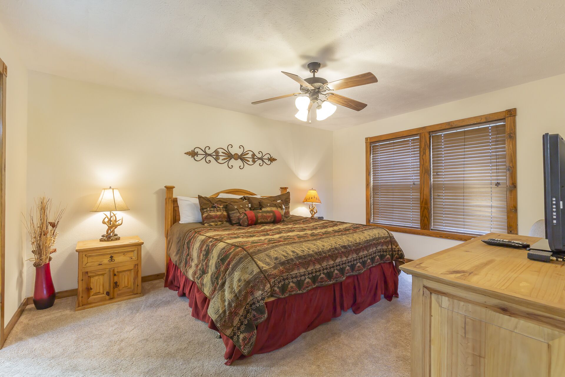 A bedroom with a bed, wooden nightstands, a ceiling fan, a TV, and a window with blinds.