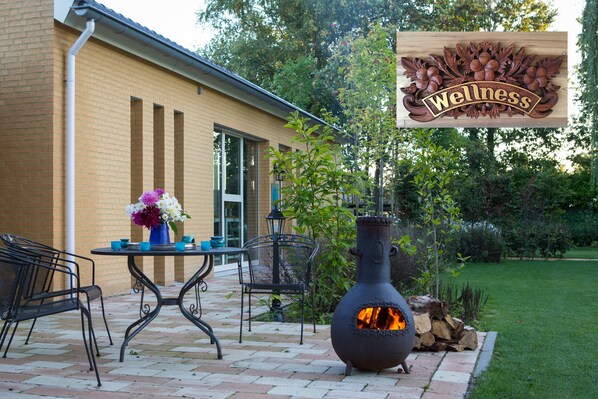 An outdoor patio with a brick floor, a metal table with chairs, a decorative vase with flowers, and a large metal fire pit with flames. - Guesthouse and free wellness in beautiful garden, close to nature and Eindhoven (Leende)