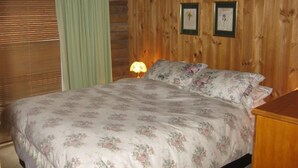 A bedroom with a wooden wall, a bed with floral bedding, a nightstand with a lamp, and two framed pictures on the wall. - Graceland Mount Victoria (Mount Victoria)