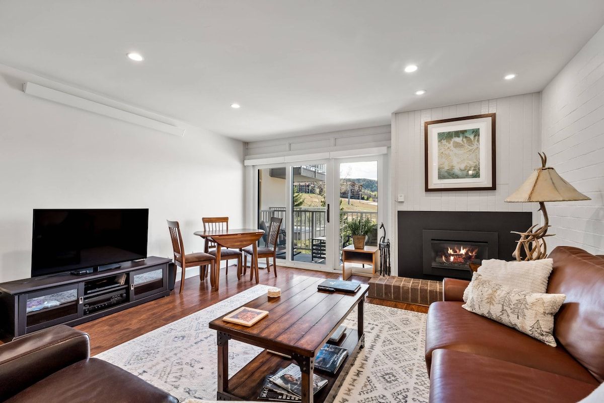 A living room with a flat-screen TV, wooden furniture, a fireplace, and a view through glass doors.