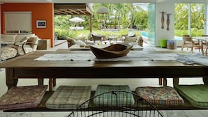 A spacious living area with a large wooden table, comfortable seating, and a view of a lush garden and pool. - Beach house with 4 suites and a pool. Comfort and privacy just steps from the sea. (Camaçari)