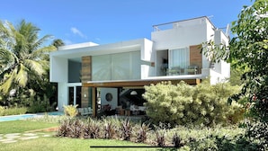 A modern two-story house with a large glass window, a pool, and lush greenery. - Beach house with 4 suites and a pool. Comfort and privacy just steps from the sea. (Camaçari)