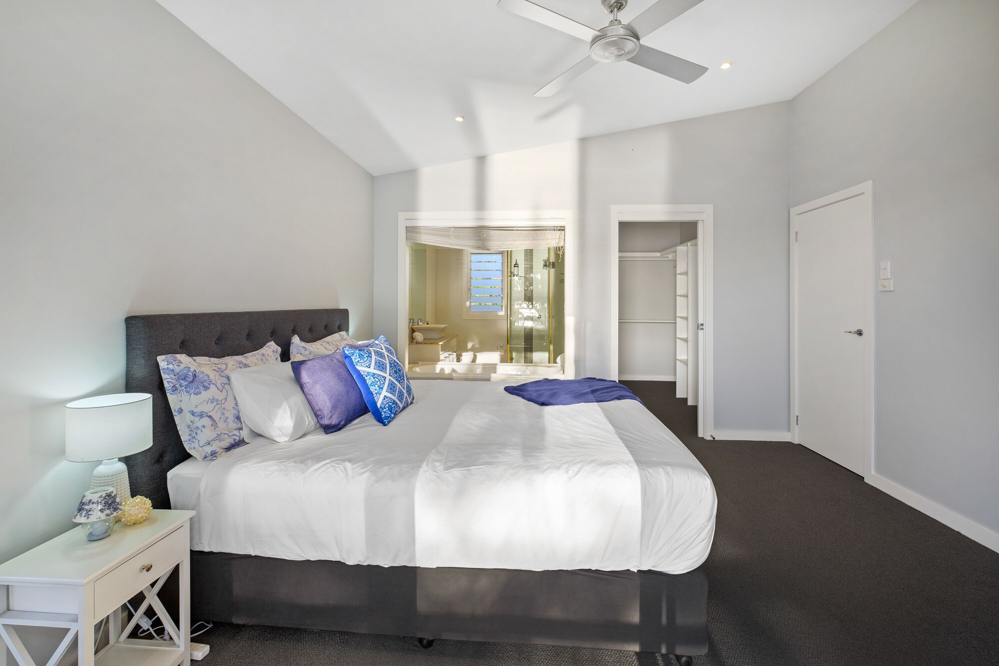 A modern bedroom with a large bed, a bedside table, a ceiling fan, and a view of the bathroom.