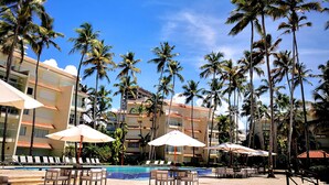 A pool area with lounge chairs and umbrellas, surrounded by tall palm trees and a multi-story building with balconies. - Beautiful Beach Front Condo, Club Hemingway, Juan Dolio, D.R (Juan Dolio)