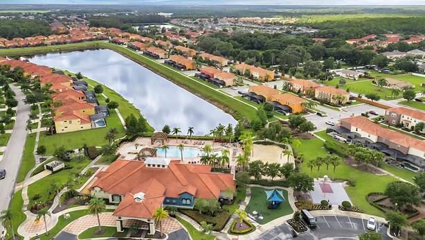Vista aérea de una zona residencial con un lago grande, casas con techos rojos y un edificio central con piscina.