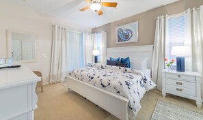 Une chambre à coucher comprenant un lit, des tables de chevet, une commode, un ventilateur de plafond et un tableau accroché au mur.