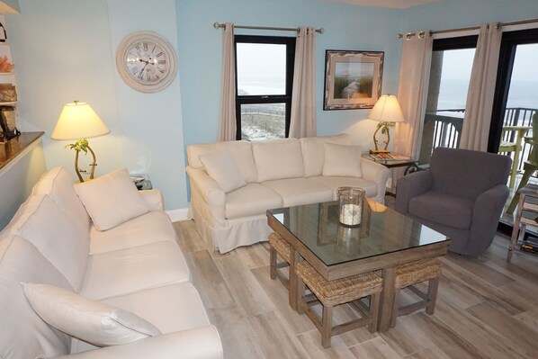 Een woonkamer met een beige bank, een glazen salontafel, een grijze fauteuil en uitzicht op de zee door grote ramen.