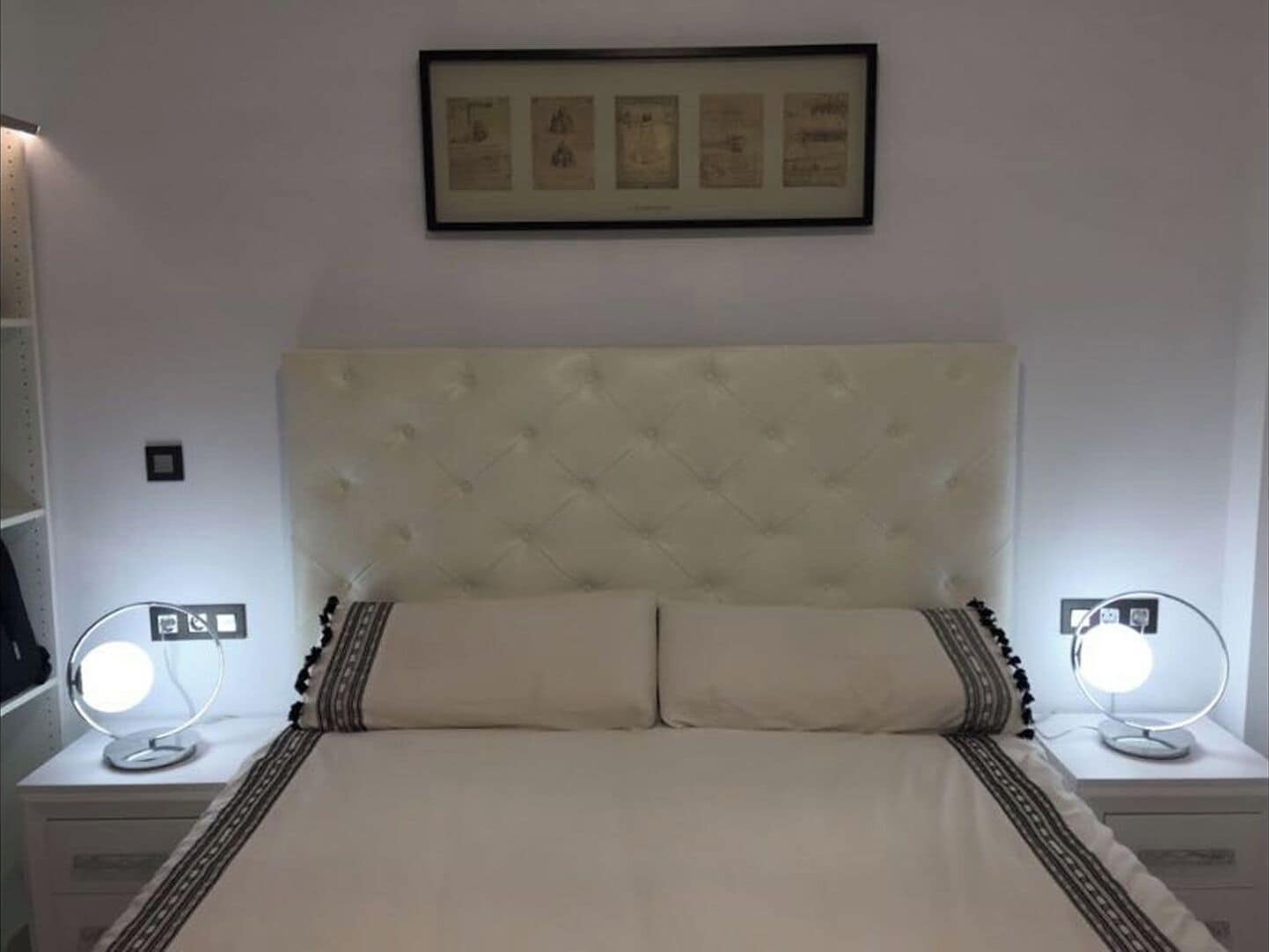 Un dormitorio con cabecera capitonada, dos mesitas de noche con espejos redondos, y un cuadro enmarcado sobre la cama.