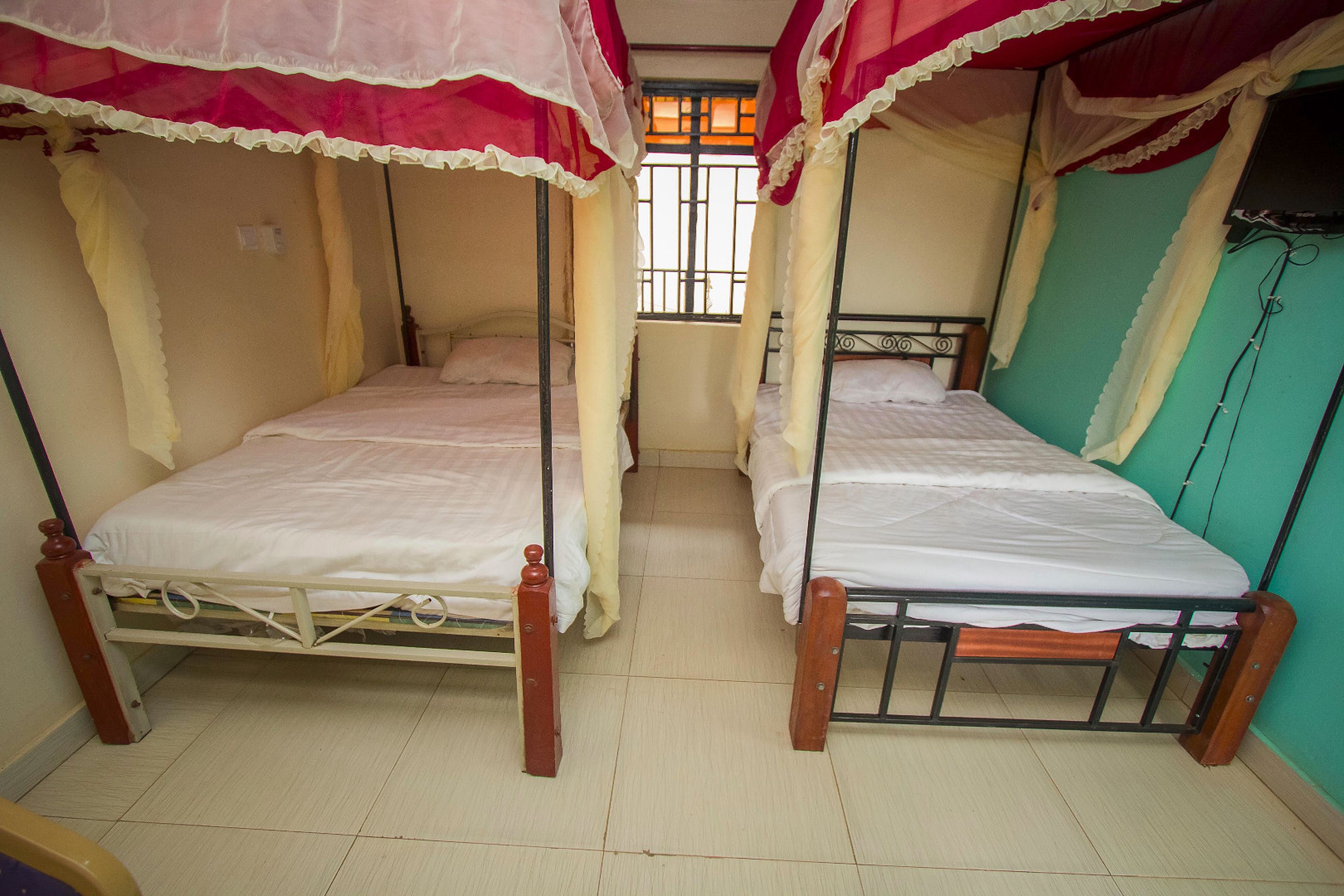 Une chambre avec deux lits simples, chacun surmonté d’un baldaquin. La chambre est dotée d’un sol carrelé et d’une fenêtre avec des barreaux.