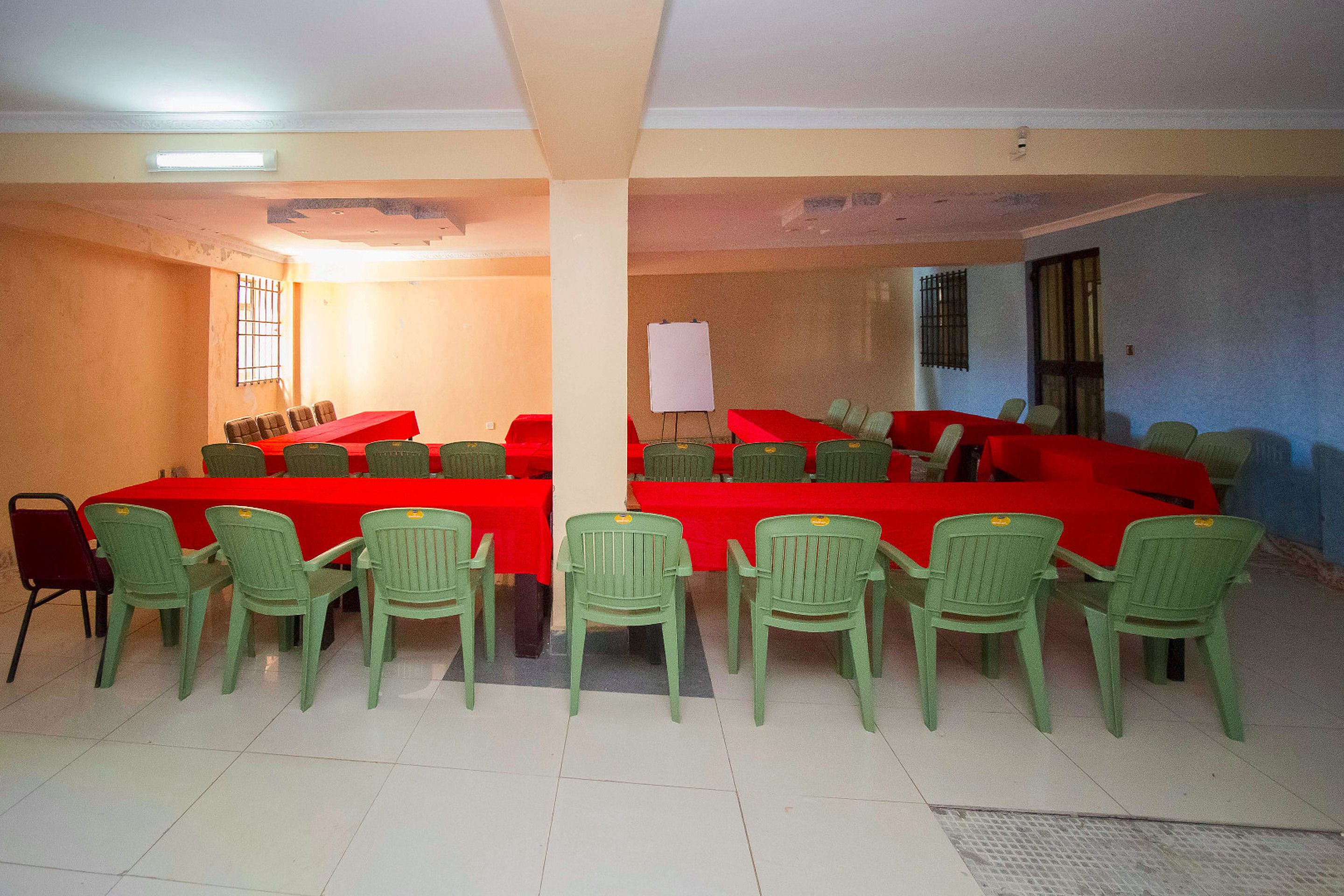 A conference room with two long tables covered with red tablecloths, surrounded by green plastic chairs.