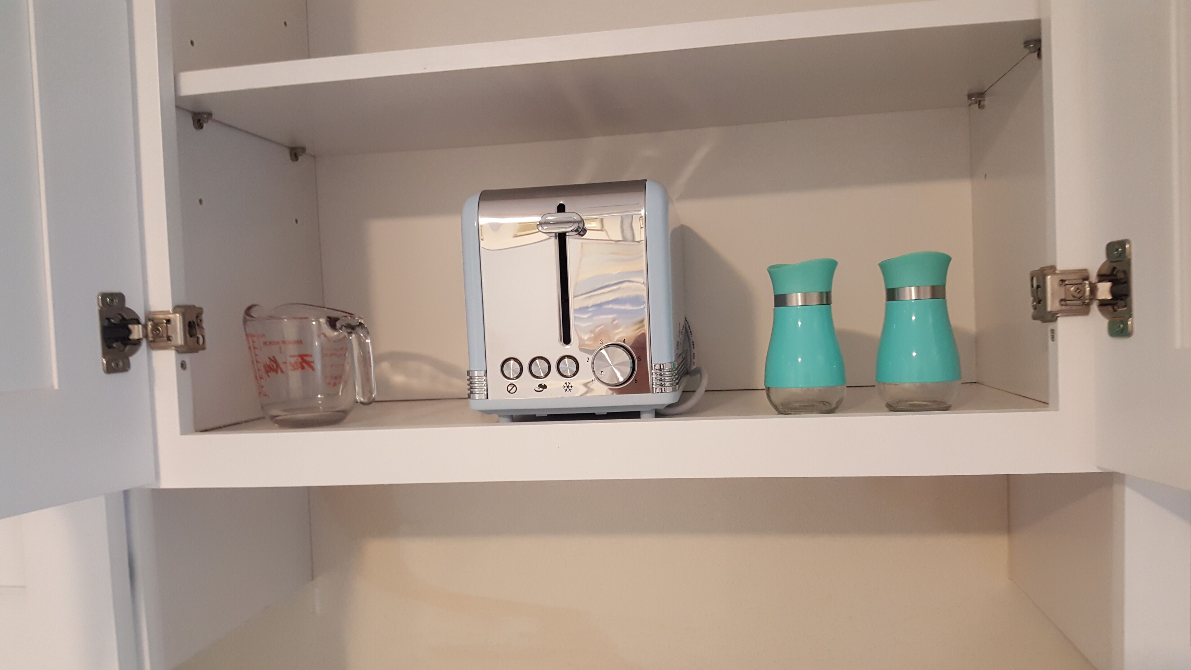 A kitchen cabinet with a toaster, a measuring cup, and two teal-colored containers.