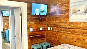 Quarto com painéis de madeira, uma cama, televisão presa à parede e porta que dá para outro espaço.