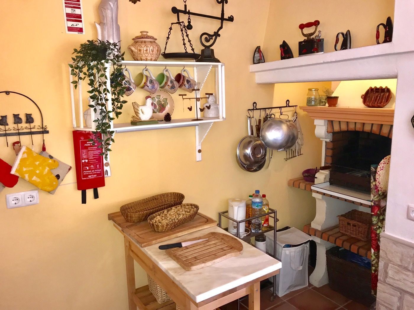 A kitchen with a brick fireplace, hanging pots, a shelf with kitchen utensils, and a red fire extinguisher.