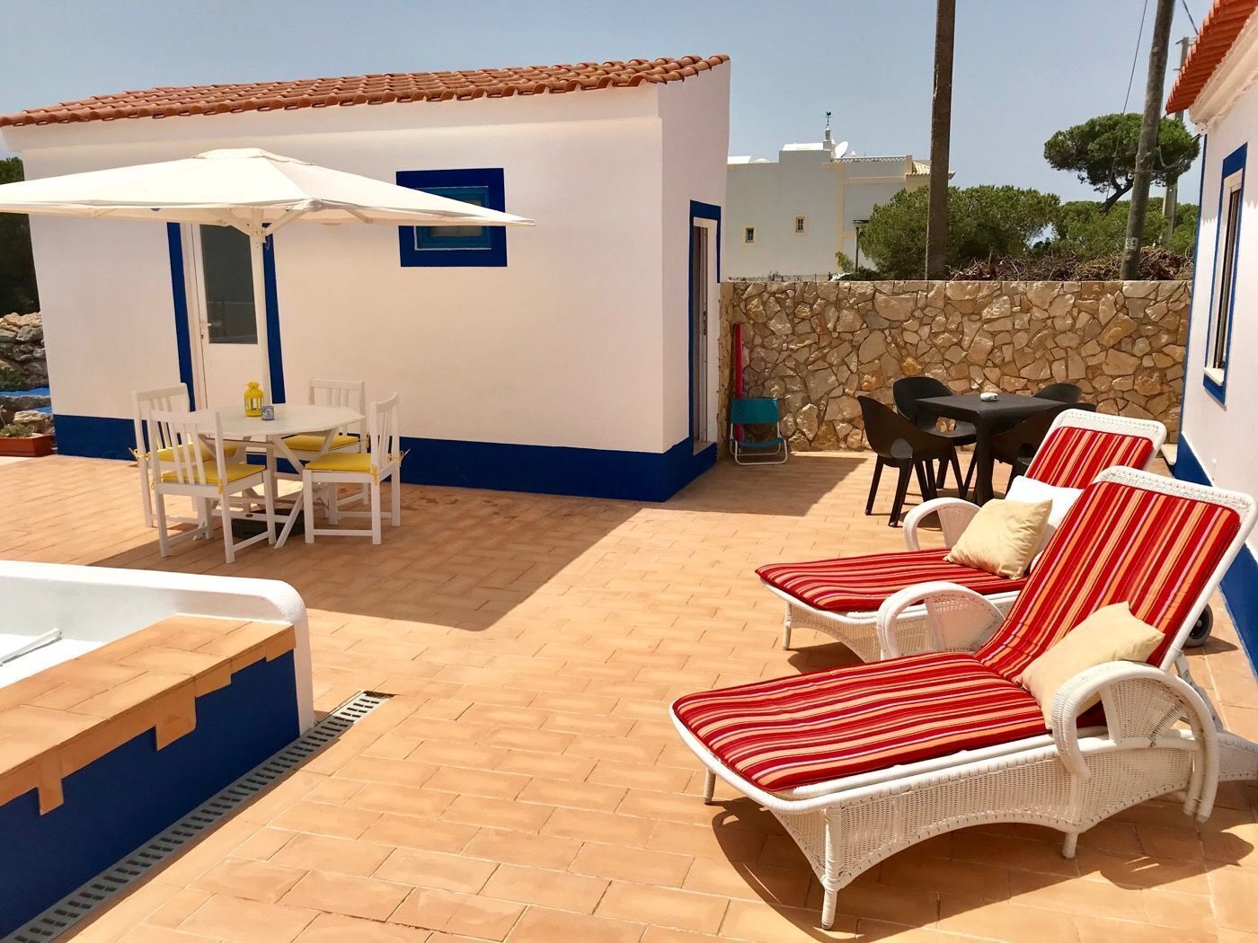 Une terrasse avec une table et des chaises blanches, un petit bâtiment blanc avec une porte bleue et un mur de pierre.