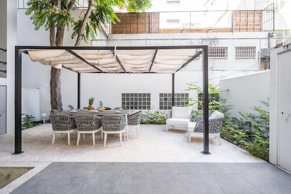 Un patio cubierto con mesa y sillas, rodeado de plantas y una cerca blanca.