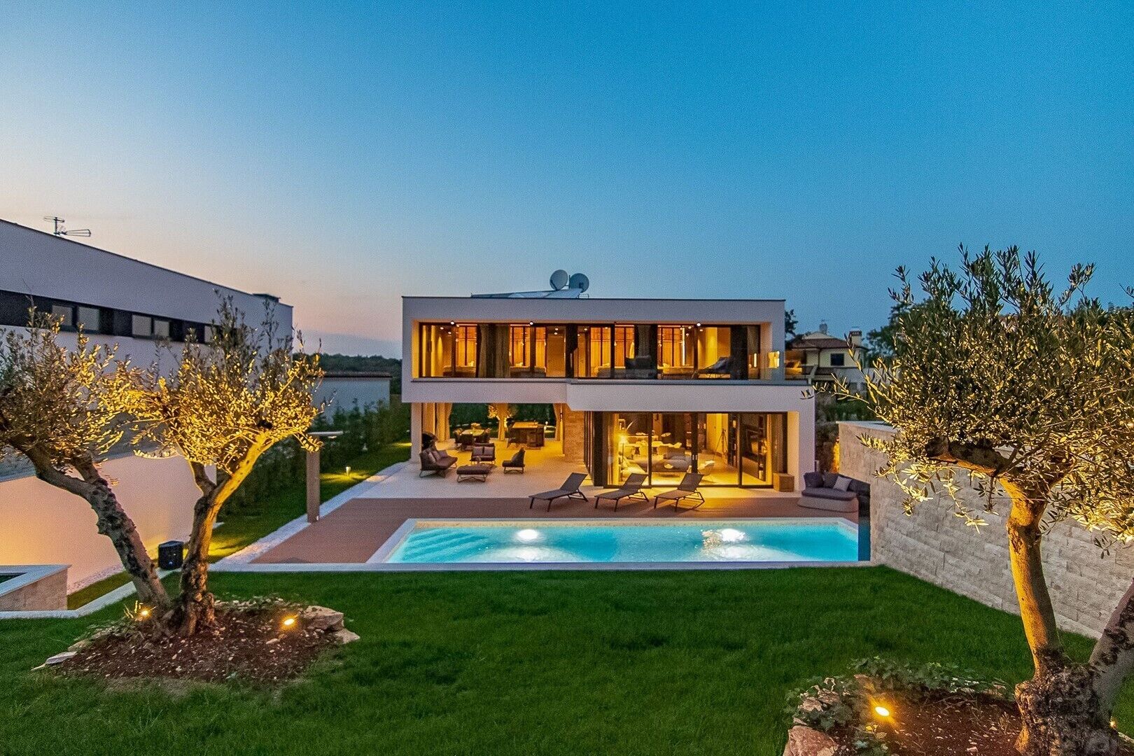 A modern two-story house with a swimming pool, outdoor seating, and well-lit surroundings.