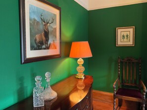 A room with a green wall, a framed deer painting, a table with a lamp, and a wooden chair.