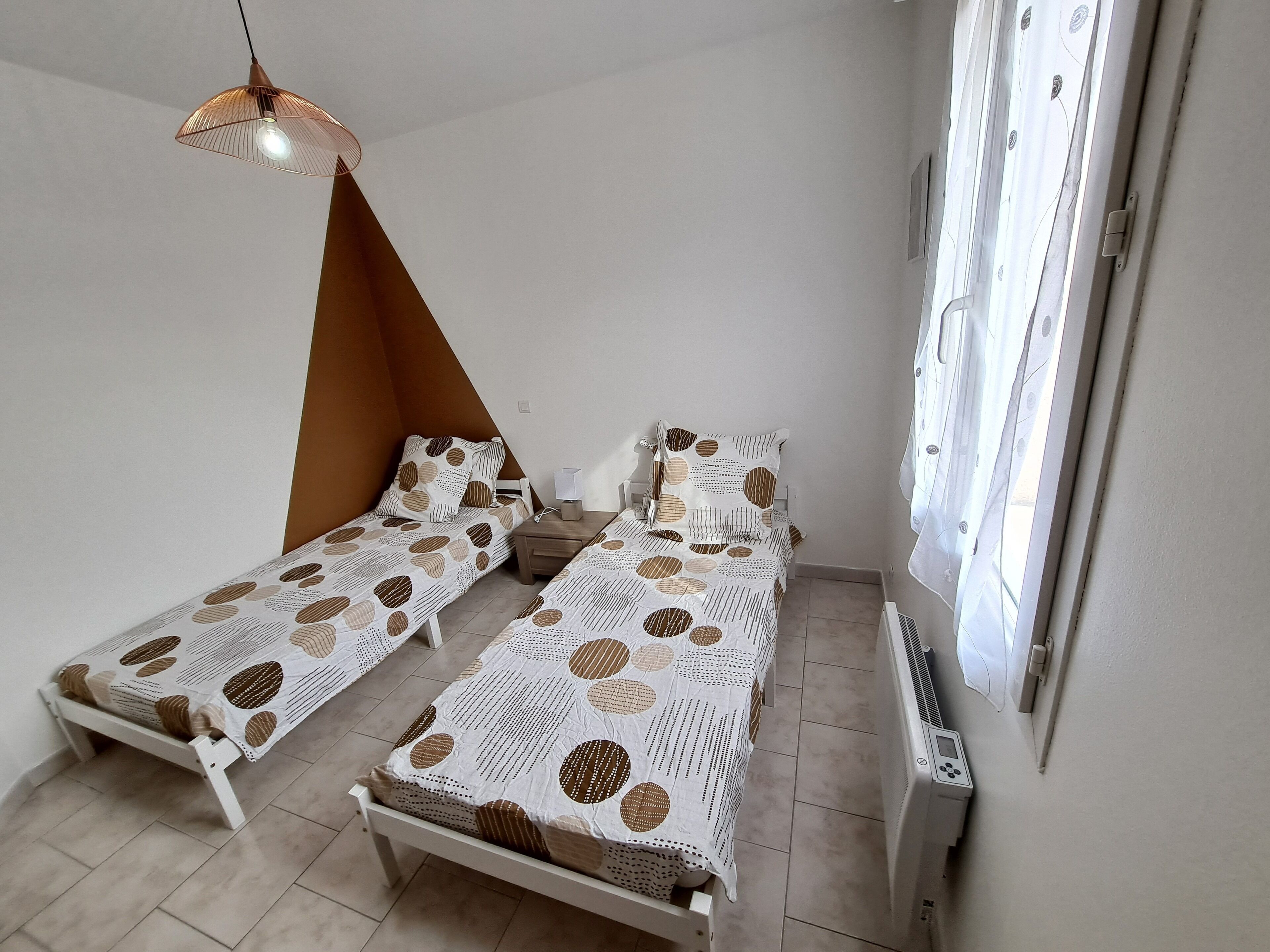 Une chambre avec deux lits simples, une table de chevet avec une lampe, un radiateur et une fenêtre avec des rideaux.