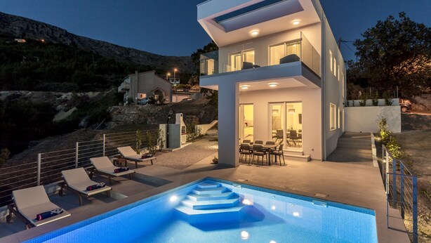 Une maison moderne à deux étages, dotée d’une piscine, d’un espace de détente extérieur et offrant une vue sur un paysage montagneux.