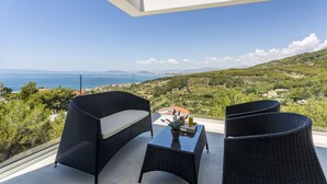 Un balcon agrémenté de meubles en osier, une petite table avec un vase, et une vue sur la mer et les collines.