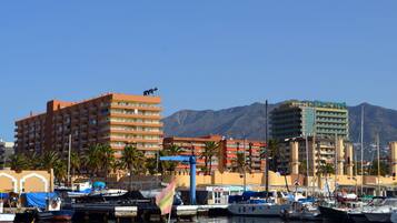 Un port de plaisance où sont amarrés de nombreux bateaux, un hôtel imposant doté de balcons, et une chaîne de montagnes en arrière-plan.