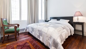 Un dormitorio con una cama grande, una silla, una mesita de noche y un ventanal con cortinas.