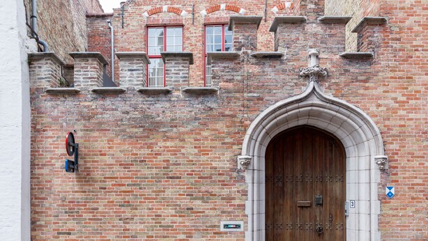 Un edificio histórico con una puerta de madera destacada, paredes de ladrillo y una pequeña ventana con un persiano rojo.