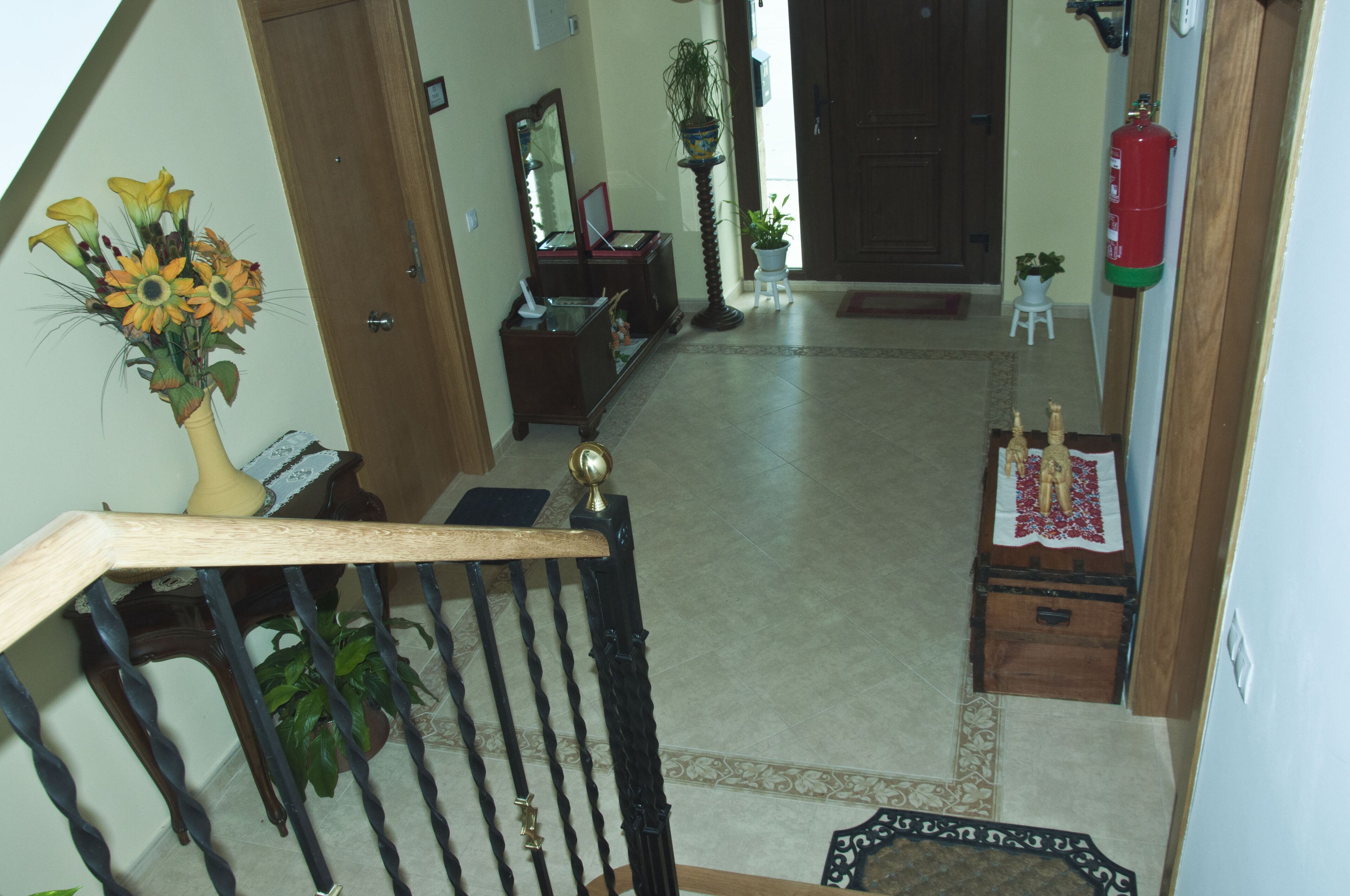Una escalera con barandilla de madera, un jarrón de flores, un espejo, una mesita con una planta y un extintor en la pared.
