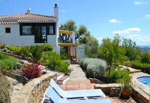 A modern house with a stone pathway leading to a pool area, surrounded by lush greenery and a clear blue sky.