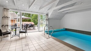 Une piscine intérieure moderne avec des parois en verre transparent, un sol carrelé et un espace extérieur aménagé avec du mobilier de jardin.