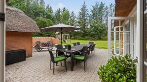 Une terrasse extérieure avec un mur en briques, une structure à toit de chaume et une table entourée de chaises.