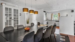 Un espace repas moderne avec une longue table, des chaises et un saladier rempli de fruits. On aperçoit, au fond, une cuisine avec des meubles blancs et un réfrigérateur.