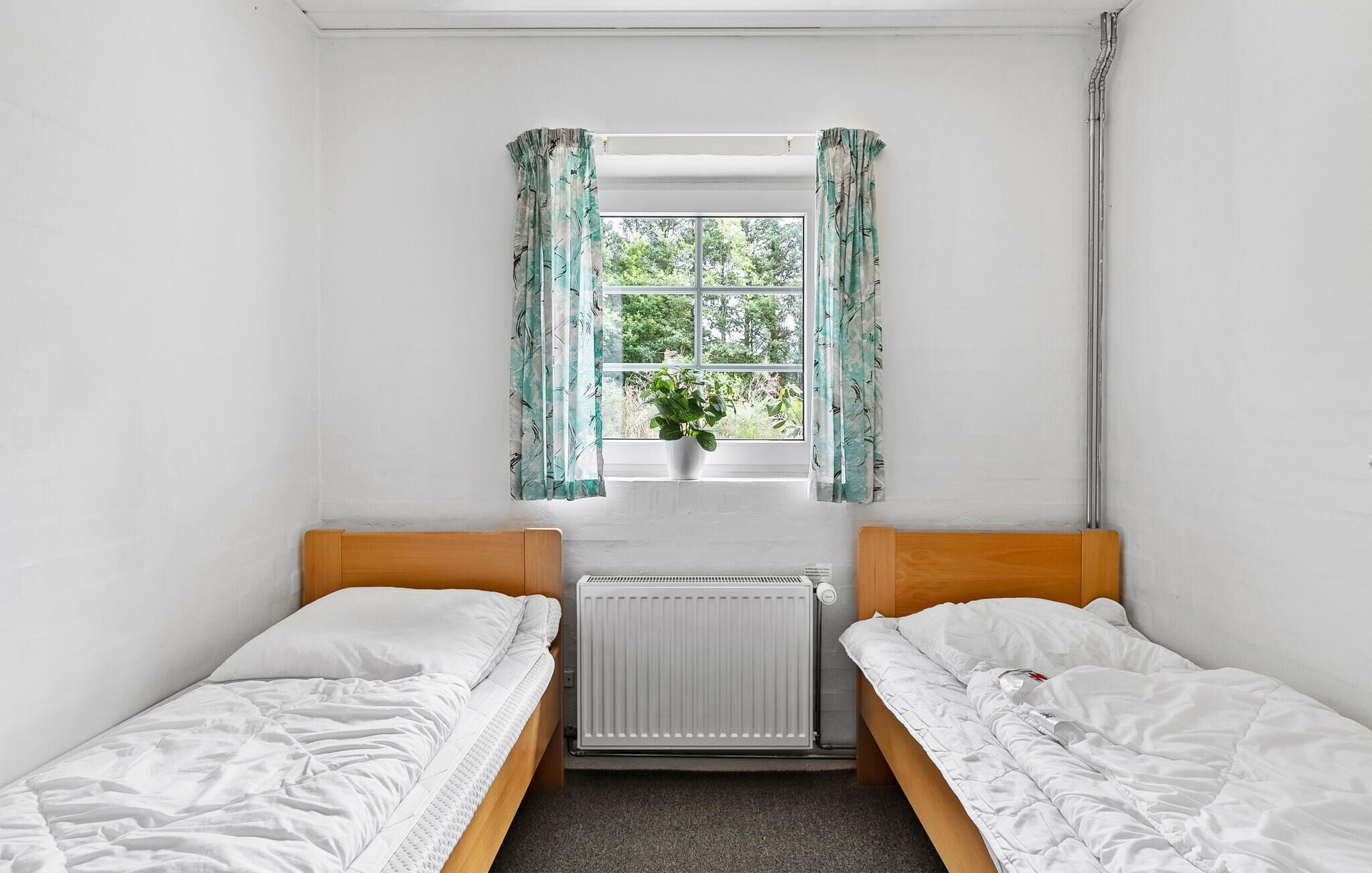 Eine kleine, einfache Unterkunft mit zwei Einzelbetten, einem Heizkörper, einem Fenster mit Vorhängen und einem Blumentopf.