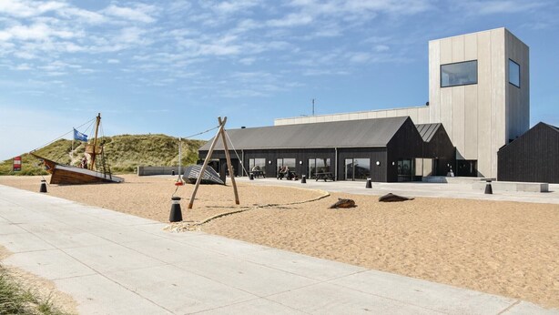 Un bâtiment moderne doté d’un toit plat et de grandes fenêtres, situé dans une zone sablonneuse, à proximité d’une promenade dallée.