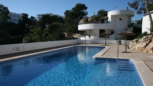 A rectangular swimming pool with clear blue water, surrounded by a white pool deck. There are umbrellas and lounge chairs around the pool area.