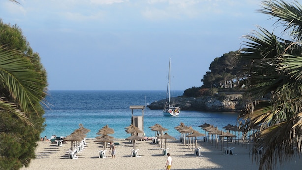 A beach with thatched umbrellas, a lifeguard tower, and a sailboat in the background.