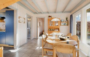 A dining area with a wooden table and chairs, a kitchen with blue cabinets, and a view of trees through large windows.
