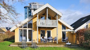 Ein zweistöckiges Holzhaus mit einem Balkon, einem Essbereich mit Gartenmöbeln und einem großen Fenster mit Blick auf den Garten.