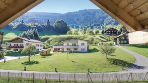Blick von einer Holzkonstruktion auf ein Dorf mit traditionellen Häusern, einer grünen Wiese und einem Berg im Hintergrund.