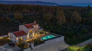 Une maison moderne à deux étages, dotée d’une piscine, entourée d’une forêt dense et avec des collines en arrière-plan.
