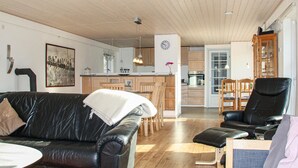 Una sala moderna con un sofá de cuero, un sillón negro y una mesita. Se ve una cocina con armarios de madera y un comedor con sillas al fondo.
