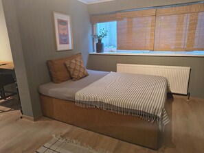 Un dormitorio con cama, un radiador, una ventana con persianas, un cuadro en la pared y una pequeña mesa con una planta.