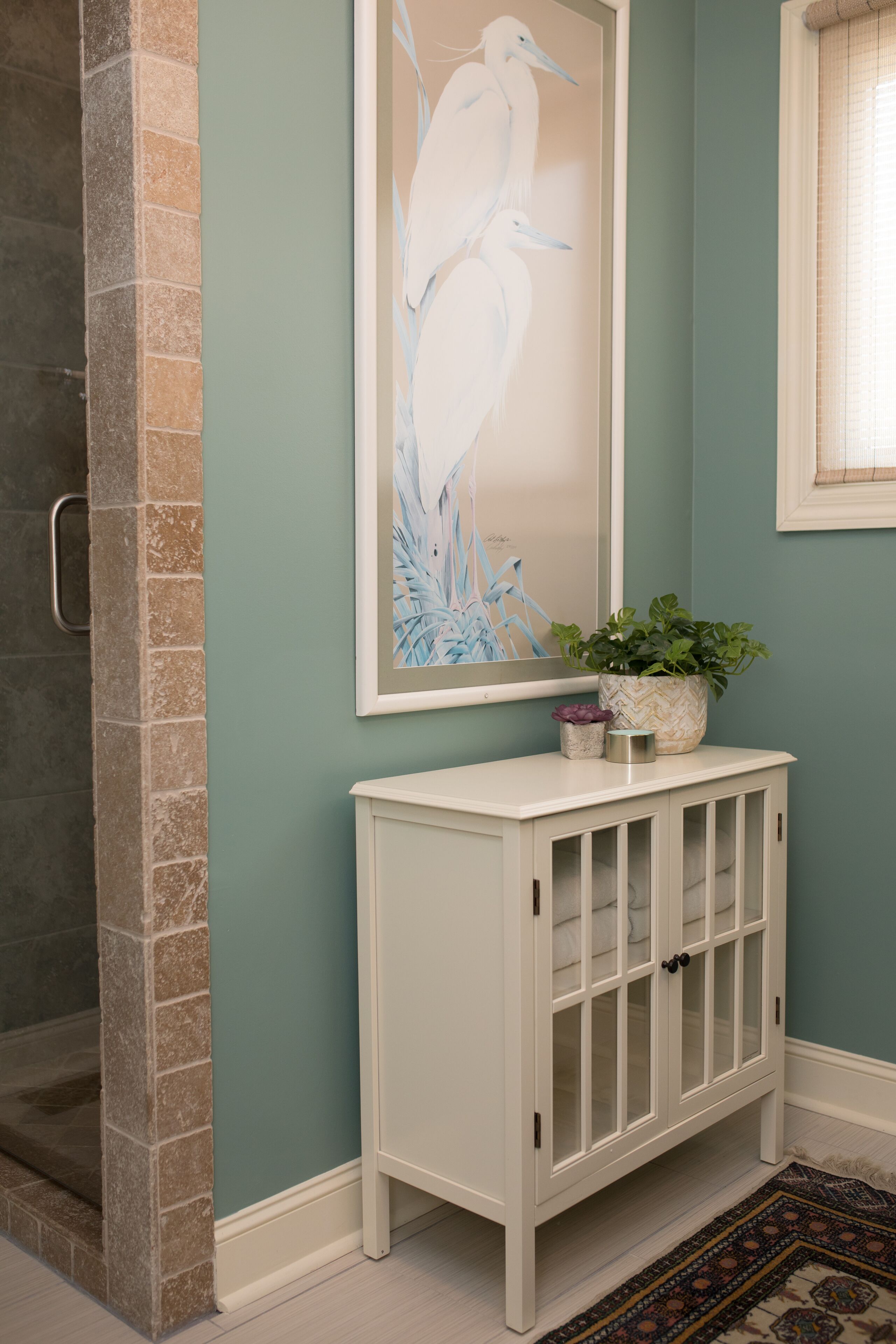 Un baño con tocador blanco, una obra de arte enmarcada con pájaros, una planta en maceta y una zona de ducha con pared de azulejos.