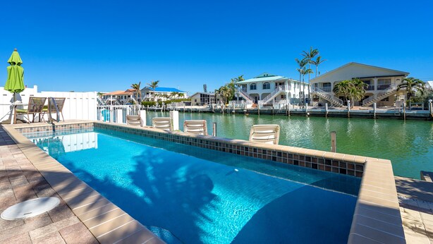 Piscina com espreguiçadeiras, um guarda-sol verde e vista para uma propriedade à beira-mar, com um edifício e palmeiras.