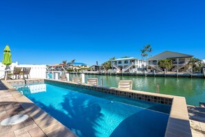 A swimming pool with lounge chairs, a green umbrella, and a view of a waterfront property with a building and palm trees. - FALL SALE ALMOST SOLD OUT 47' DOCK, PRIVATE POOL, CABANA BEACH CLUB 4 MIN WALK (Key Colony Beach)