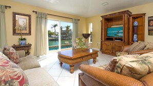 Uma sala de estar ampla com uma televisão grande, assentos confortáveis, uma mesa de centro de madeira e vista para uma piscina através de grandes portas deslizantes.