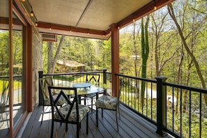 A covered outdoor deck with a table and chairs, surrounded by a wooden railing and overlooking a forested area.