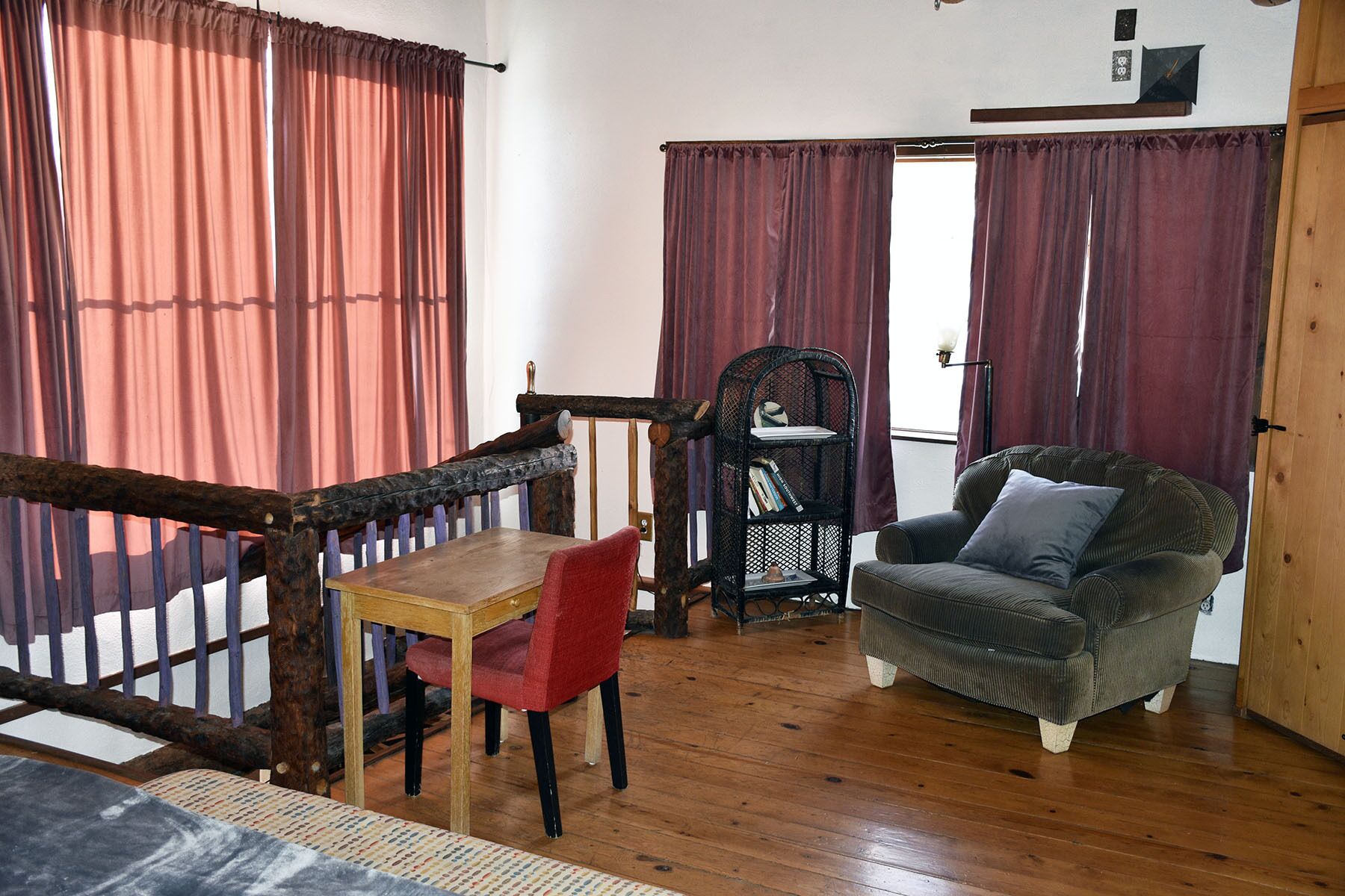 A cozy room with a wooden floor, a red chair, a green sofa, and a small table. There is a window with curtains, a door, and a shelf with books.